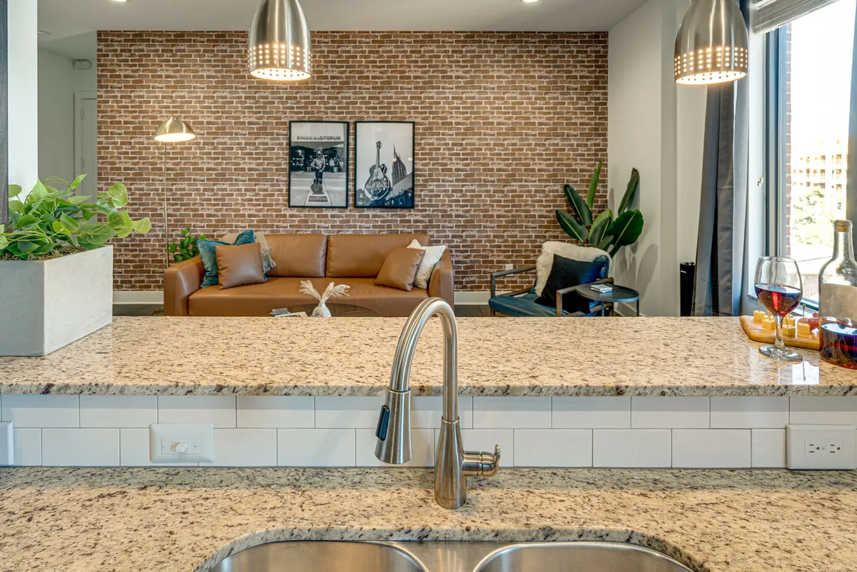 A stylish living area featuring a granite countertop in the foreground, a chic sofa (potentially a sleeper sofa), decorative plants, a brick accent wall, and vibrant artwork, creating a welcoming space for relaxation or hosting guests.