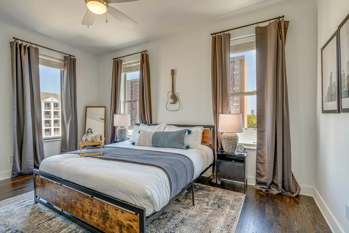 A bright bedroom featuring a plush king-size bed adorned with soft bedding, large windows providing natural light, a guitar leaning casually in the corner, nightstands with lamps, and a cozy inviting atmosphere.