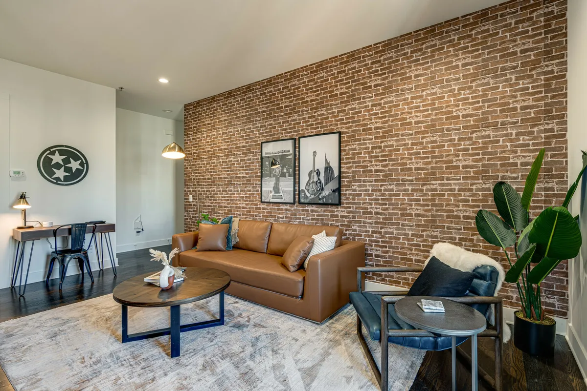 A stylish living room featuring an exposed brick wall, a plush leather sofa, a coffee table, modern artwork, and a vibrant green plant, all illuminated by abundant natural light pouring in through large windows. The space blends industrial textures with soft, modern furnishings, offering a welcoming and relaxing environment.