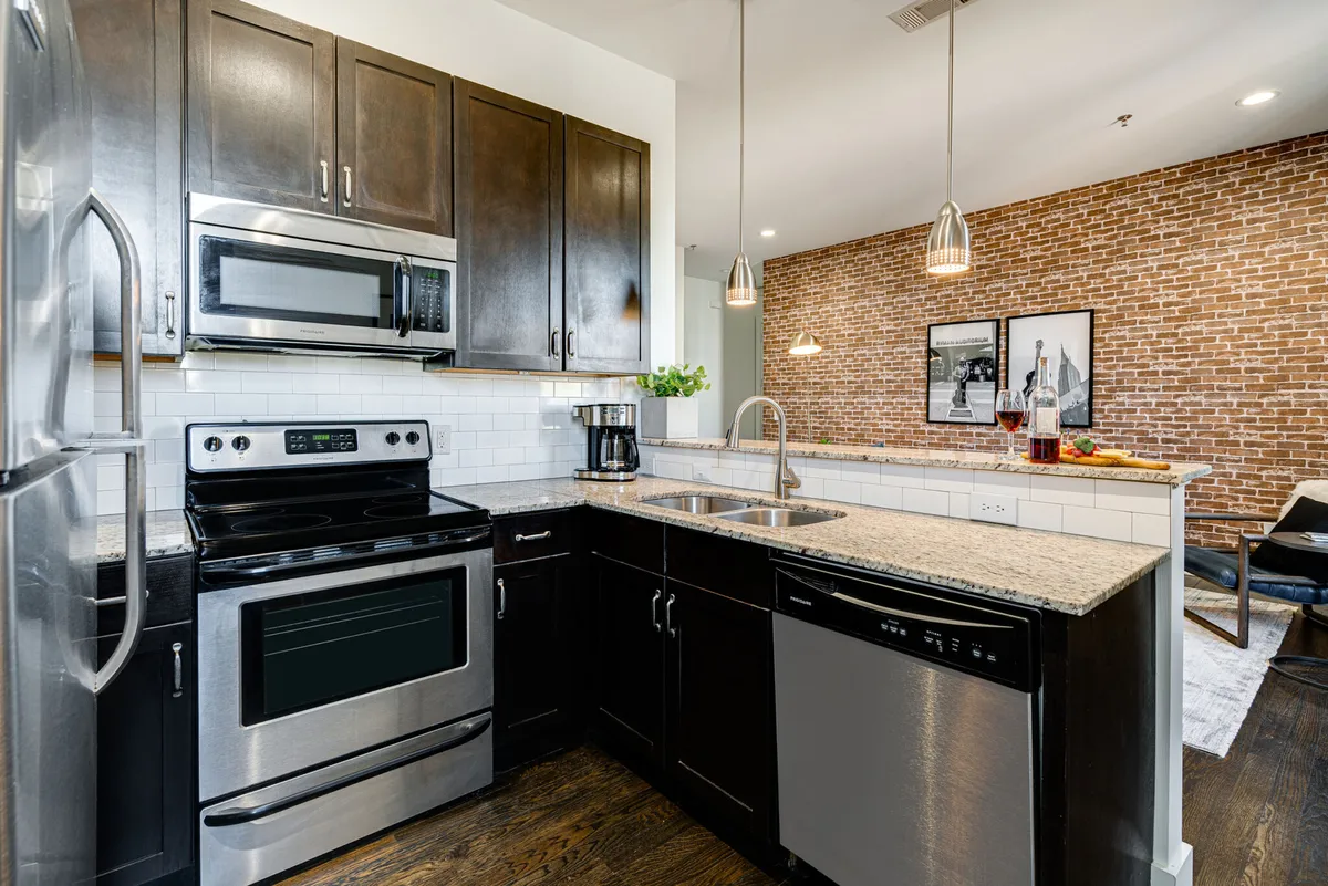 A stylish kitchen featuring granite countertops, sleek stainless steel appliances, exposed brick walls, and chic pendant lighting. The scene is set up for cooking and entertaining, with an inviting atmosphere that feels both modern and welcoming.