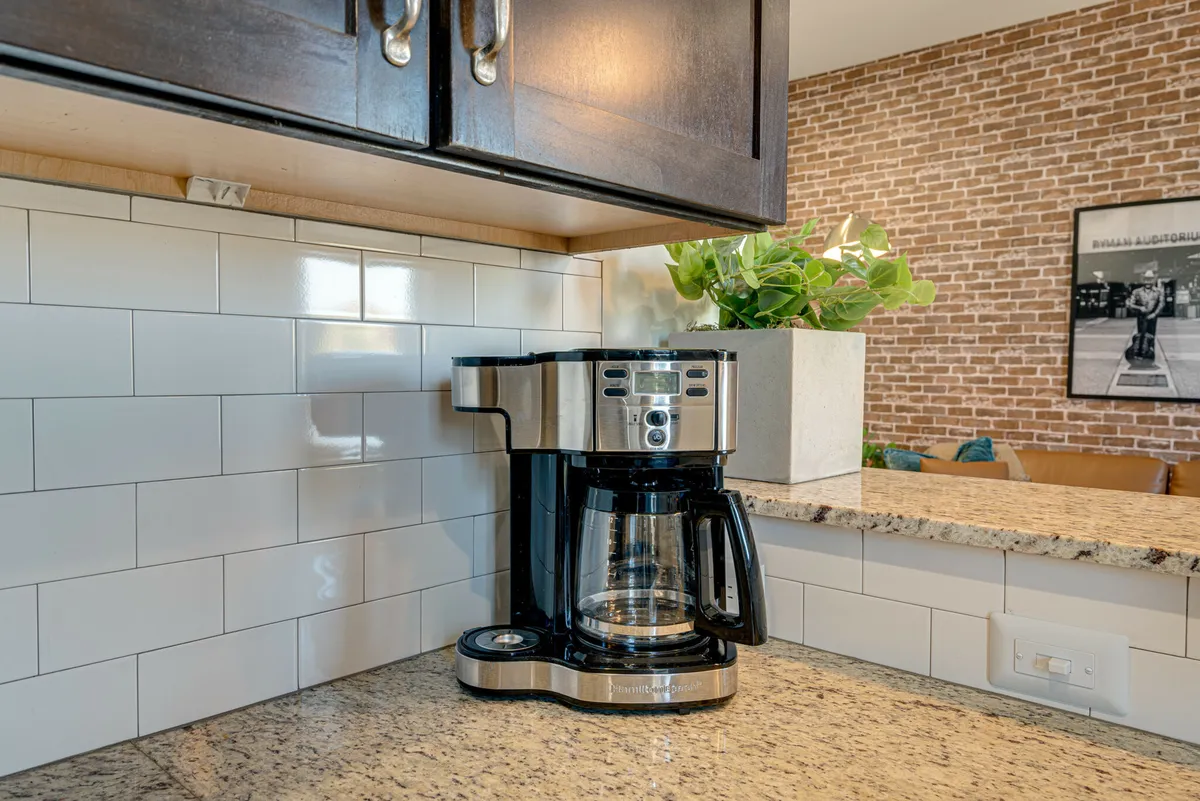 A cozy kitchen corner featuring a coffee maker on a granite countertop, a brick accent wall, and a small plant, suggesting a perfect spot for morning coffee or a late-night snack.