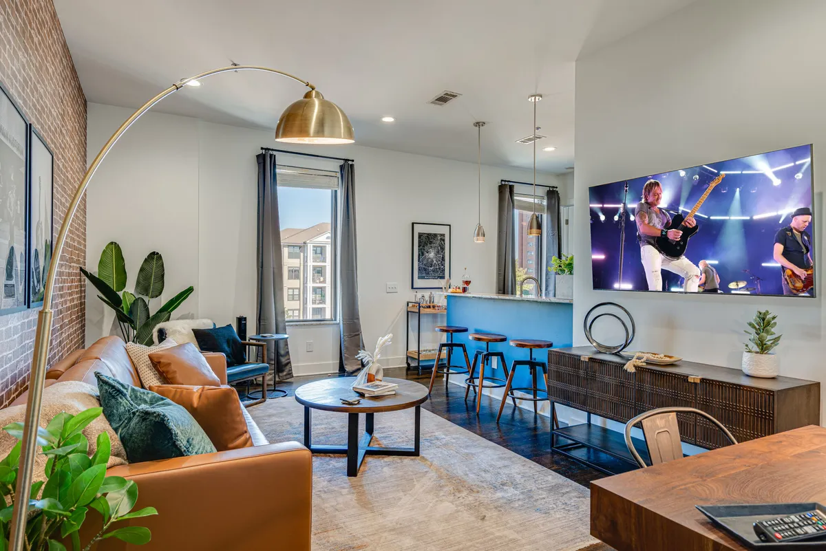 The image showcases a sunlit, open-concept living area featuring a plush sofa, wall-mounted television, and a cozy dining nook accented by lush green plants. The space feels comfortable and lively, inviting guests to relax, entertain, or unwind with views of the city beyond.