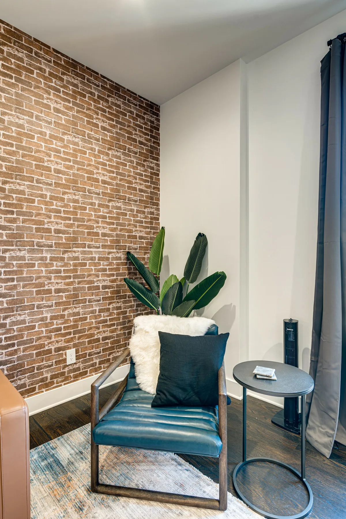 A modern, comfortable interior featuring a brick wall, blue accent chair, green plant, and a small side table. The room is bathed in natural light, creating an inviting atmosphere for relaxation or socializing.