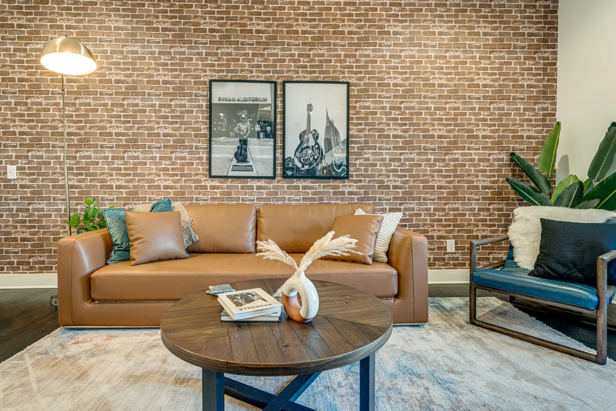 A stylish living room setup featuring a brown leather sofa, round wooden coffee table, framed wall art, and green plants. The room is bathed in daylight, creating a cozy nook ideal for reading, socializing, or simply unwinding above city life.