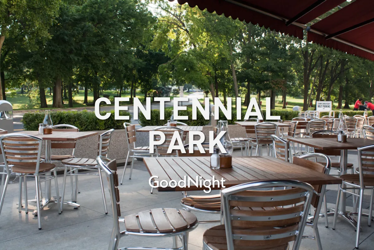 A relaxed outdoor dining area with wooden tables, metal chairs, leafy green trees, and a winding pathway. The scene invites guests to imagine sunset dinners, socializing with friends, or enjoying a casual brunch in a park-like urban setting.