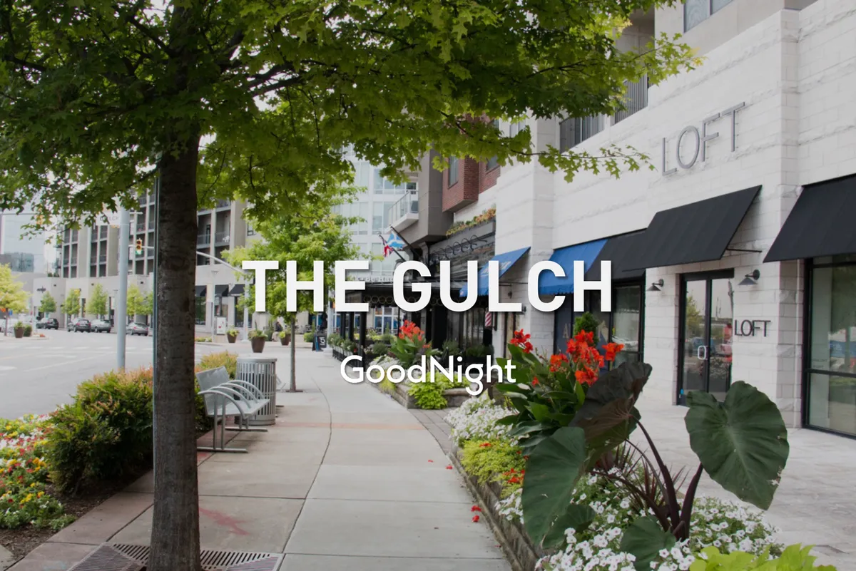 A vibrant exterior streetscape featuring lush trees, colorful flowers, charming storefronts, and benches lining a wide sidewalk. People are seen shopping, walking, and socializing, creating a lively yet family-friendly urban atmosphere ideal for morning strolls.