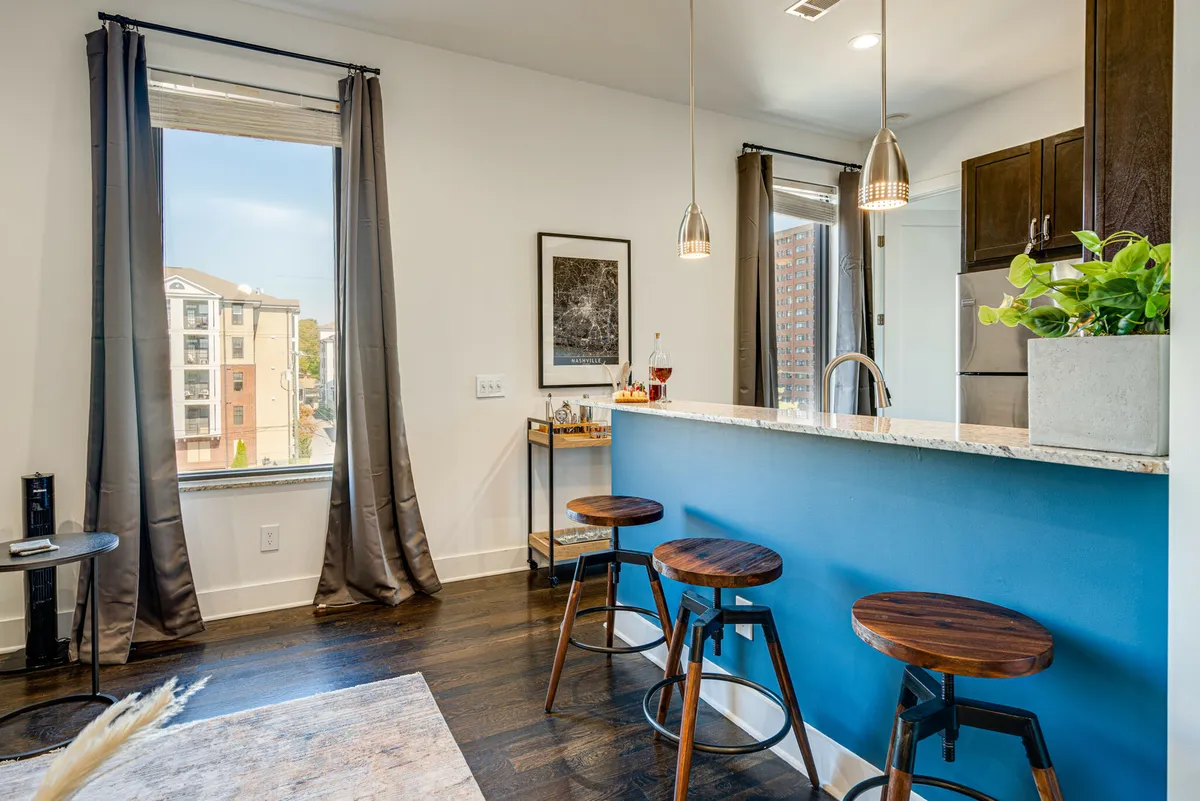 A bright, sunlit kitchen and dining area with a central island, bar stools for casual dining, a window view, curated artwork, and a touch of greenery from a potted plant. The open concept encourages socializing and connection.